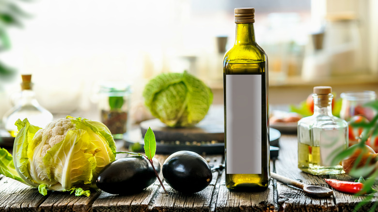 A dark glass bottle of olive oil rests on a wooden surface near vegetables and other bottles