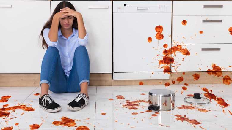 A person sits on a dirty kitchen floor next to a spilled pot of red sauce
