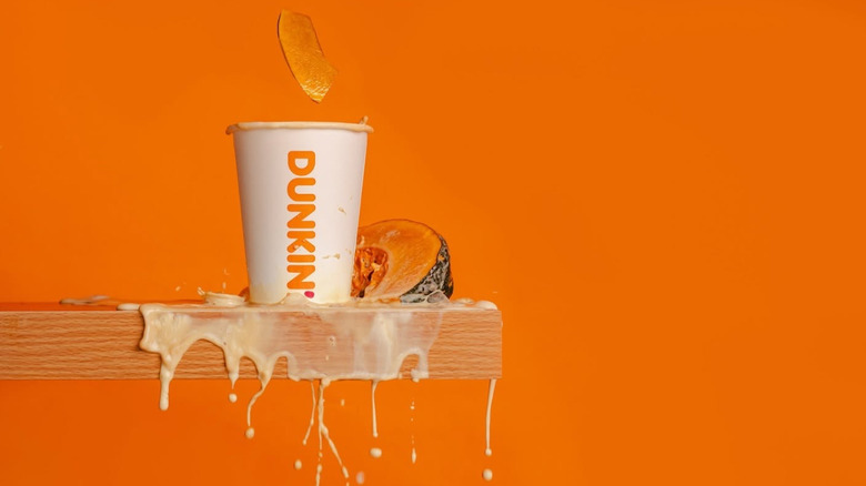 A cup of Pumpkin Spice Latte from Dunkin' overflows as chunks of pumpkin fall in.