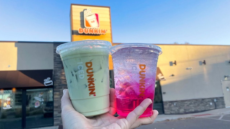 Hand holding Wicked Pink and Wicked Green Matcha Refreshers in front of Dunkin' storefront