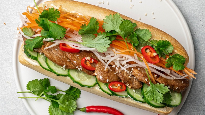 An open-faced bánh mì sandwich sits on a white plate next to herbs and chiles