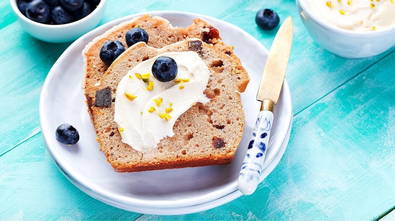 Two slices of banana bread on a plate topped with a smear of cream cheese frosting, chopped pistachios, and whole blueberries