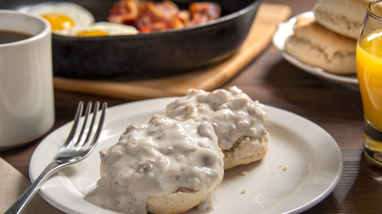 Biscuits and gravy on a white plate with juice, coffee, eggs, and bacon in the background
