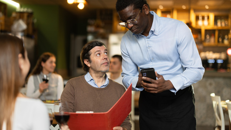A man in a restaurant holds a red menu as a waiter stands over him waiting to take his order
