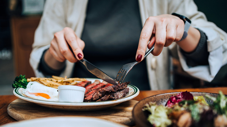 Woman cutting into steak on a white plate with a sunny-side up egg, fries, and broccoli