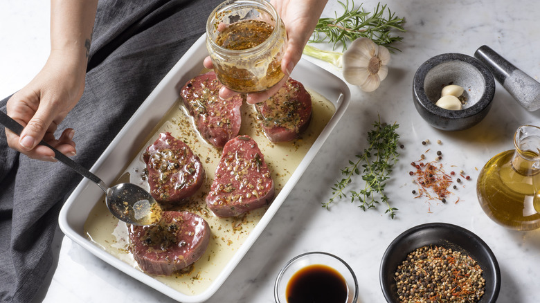 Person's hand dropping marinade over raw steaks in a white dish surrounded by spices, herbs, sauce, and garlic