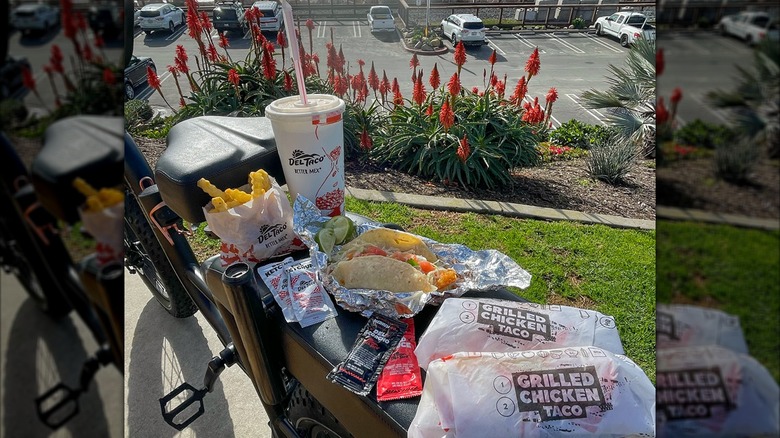 A feast of food. including tacos, crinkle cut fries, hot sauce, and a drink, from Del Taco on a bicycle at the beach.