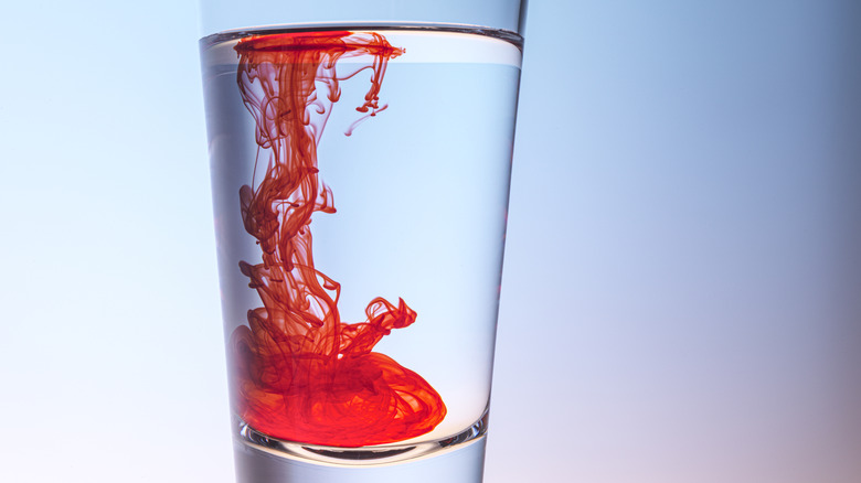 A shot glass contains red food dye floating in water against a light blue backdrop