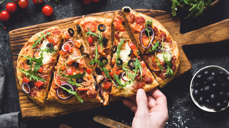 A person's hand reaches for a slice of flatbread pizza topped with cheese, olives, tomatoes, and meat