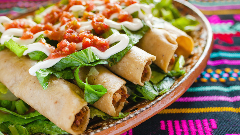 A plate of flautas topped with lettuce, sour cream, and salsa sits on top of a Southwestern-patterned fabric