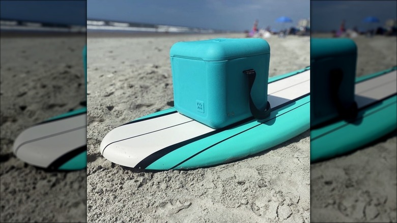 A turquoise FOAM cooler sits on a surfboard on a sandy beach