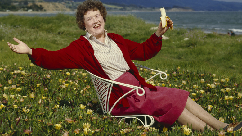 Julia Child, smiling broadly with her arms spread out, sits in a field near the seaside, holding a partially eaten banana