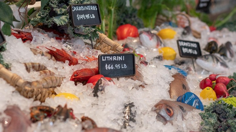 various fish, shellfish, and vegetables on ice with chalkboard signs indicating the fish and price