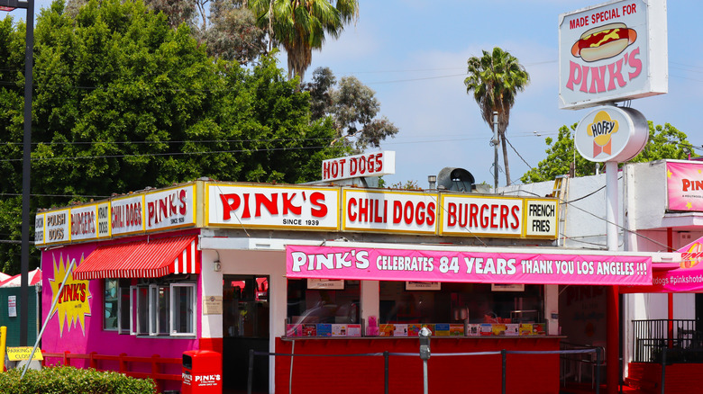 exterior of pink's hot dog stand with colorful signs, awnings, and banners in front of trees