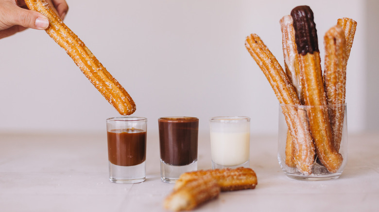 A person dipping a churro in three kinds of sauce (dulce de leche, chocolate, and condensed milk) next to a glass filled with churros