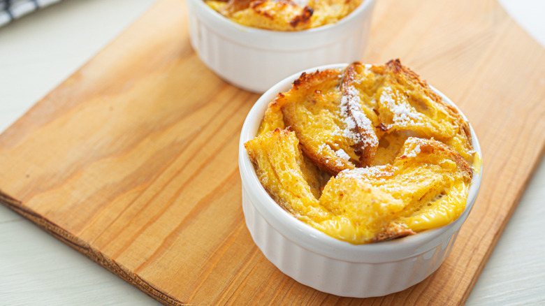 Bread pudding in a white ramekin on a wooden cutting board, a second bread pudding in the background