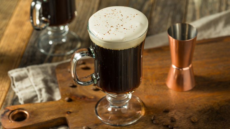 Coffee with a layer of whipped cream on top in a tall clear glass with a handle on a wooden surface