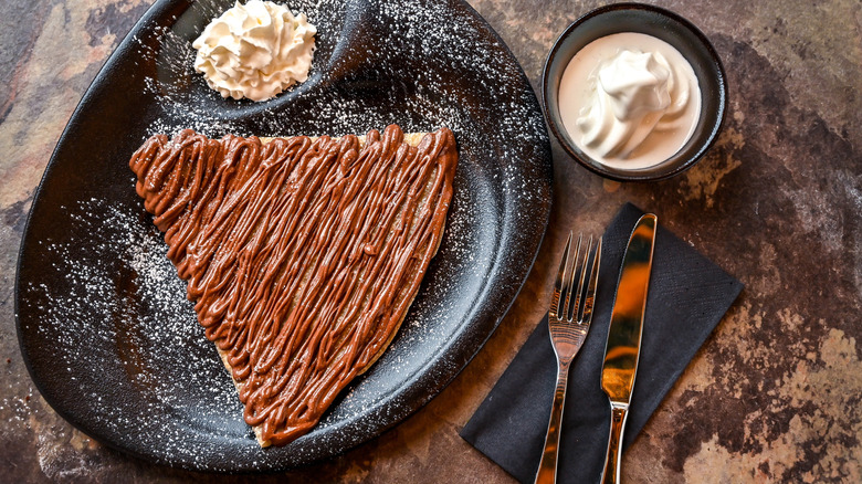 Crepe covered in Nutella on a plate with whipped cream, beside it a bowl of ice cream and cutlery