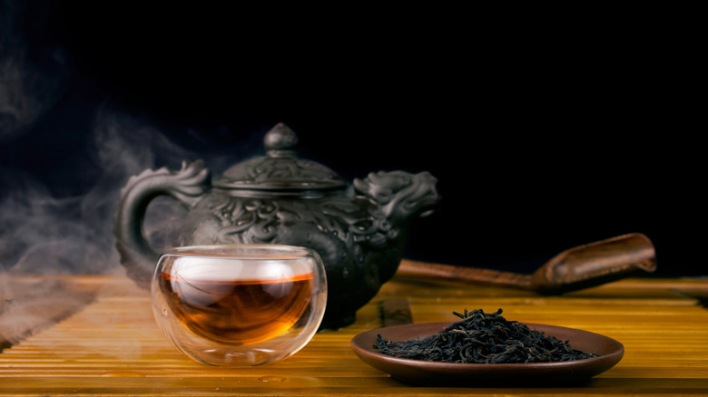 Clay teapot with red tea Lapsang souchong on a black background, heap of dry tea leaves and glass thermo cup of hot tea with vapour
