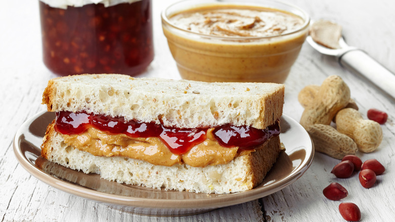 A fresh, thick peanut butter and jelly sandwich on a plate with peanut butter and jelly in the background and peanuts next to it