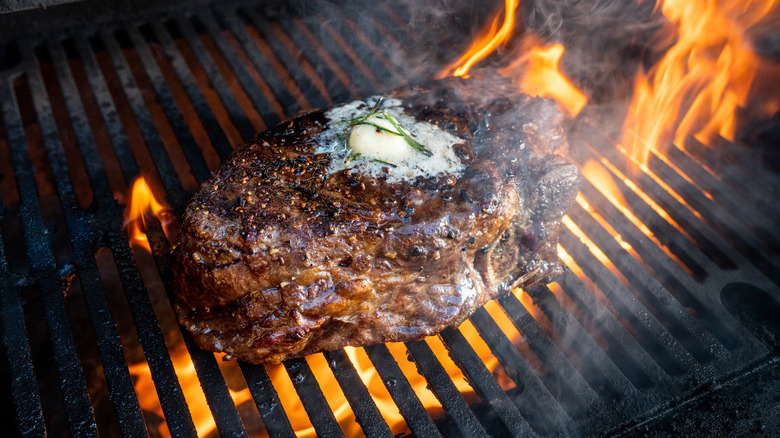 A ribeye steak grilled over an open flame has rosemary and a pat of butter melting on top.