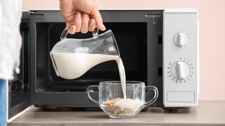 A hand pours milk into a glass mug with oatmeal, outside a microwave