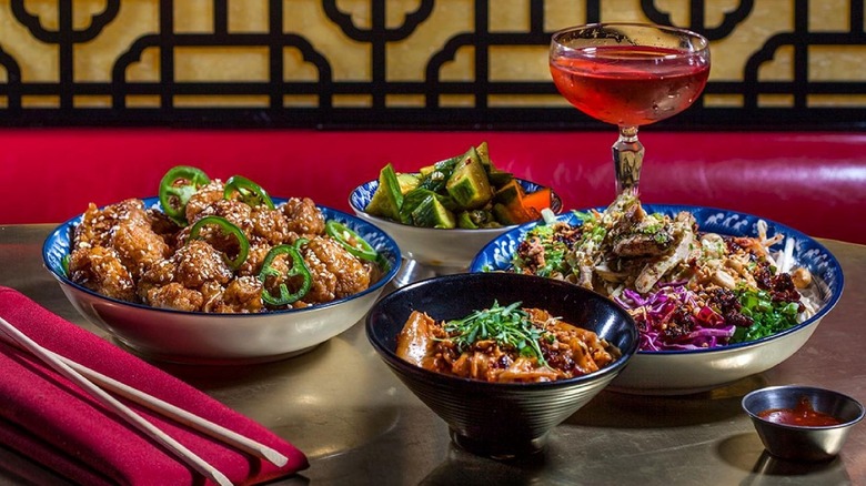 Four bowls of different Chinese foods and a pink cocktail on a table with chopsticks
