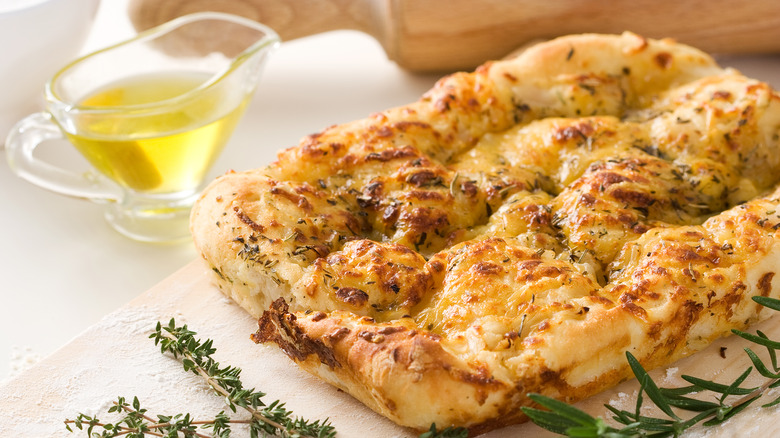 A rectangular loaf of focaccia topped with herbs and a jug of oil on the side