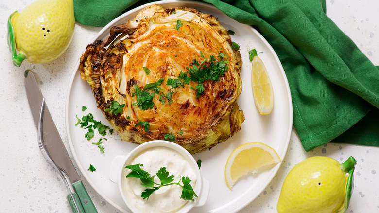 A grilled cabbage steak sits on a white plate next to lemon wedges and a small bowl of sour cream