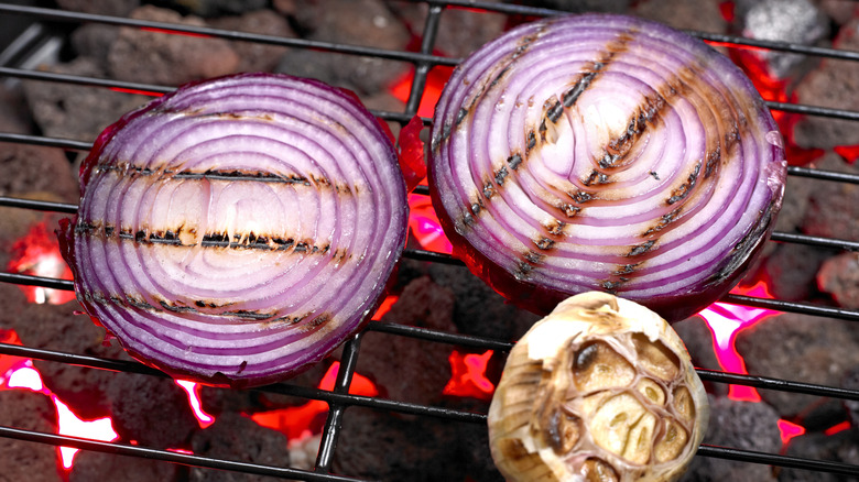 Slices of red onion sit on a grill grate over glowing charcoal bricks beside a head of garlic