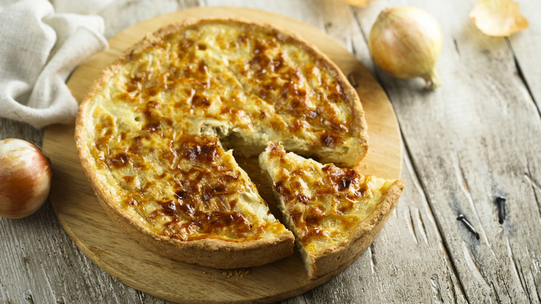 A quiche on a serving board, on a table, with some onions