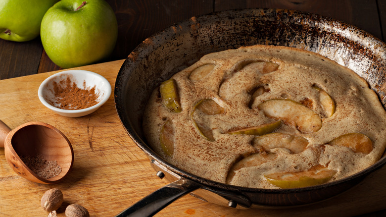 An apple pancake in a pan next to a bowl of cinnamon and nutmeg