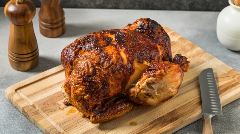 A freshly cooked rotisserie chicken sits on a wooden cutting board with a knife