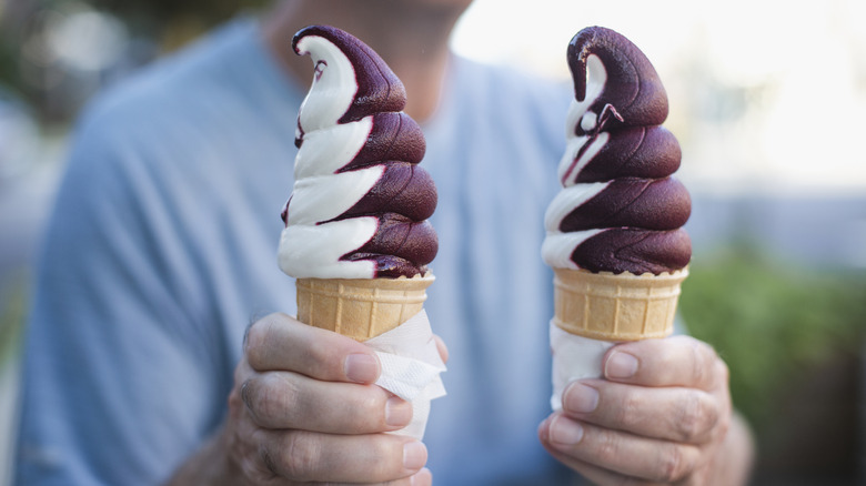 Two vanilla and berry swirl frozen yogurts in a waffle cone are held by a man wearing a light blue shirt