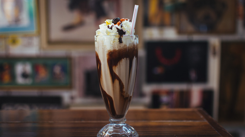 A chocolate milkshake with whipped cream and M&Ms on top sits in a glass cup on a wooden table with a plastic straw