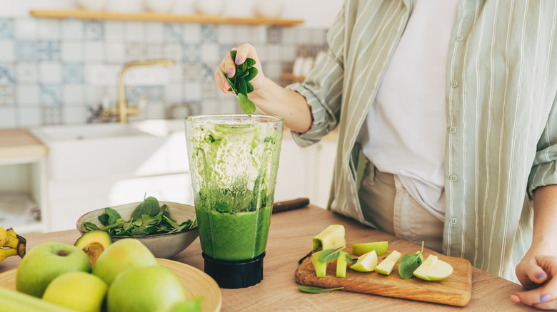 Someone prepares a green smoothie, adding spinach and apples to a blender