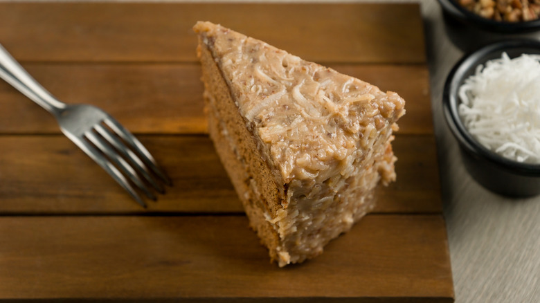 A slice of chocolate cake on a cutting board next to a fork and ramekins of shredded coconut and pecans