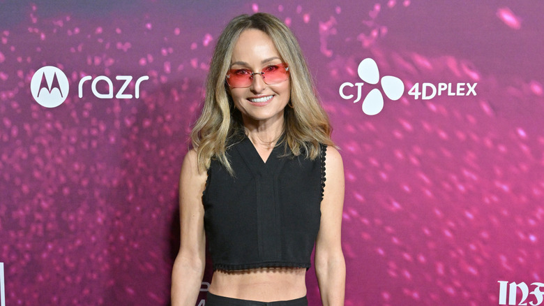 Giada De Laurentiis stands on a red carpet, smiling, and wearing rose-colored sunglasses.