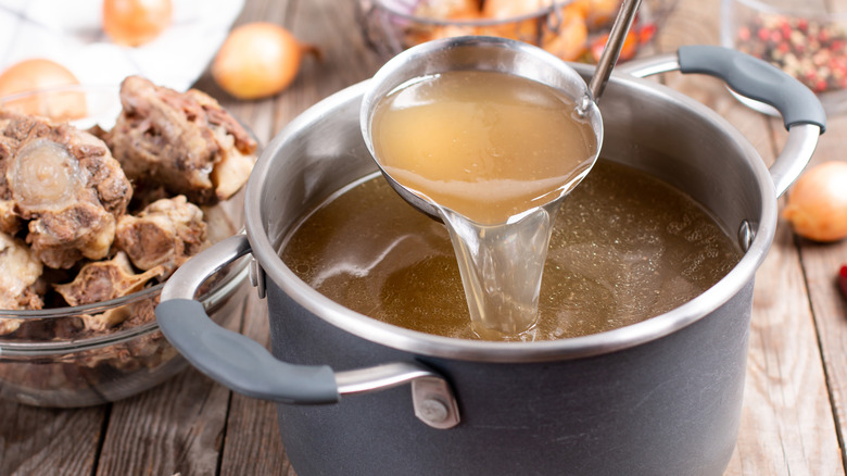 A metal ladle is dipped into a pot of beef broth, with remnants of its ingredients in the background