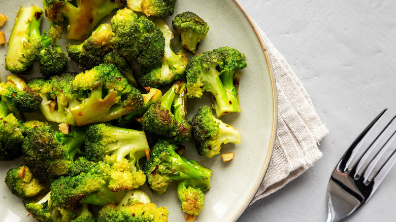 A plate of slightly browned, seasoned broccoli on top of a napkin next to a fork