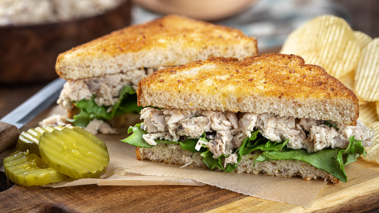 Tuna salad sandwich on toasted whole grain bread with potato chips and pickles on a cutting board