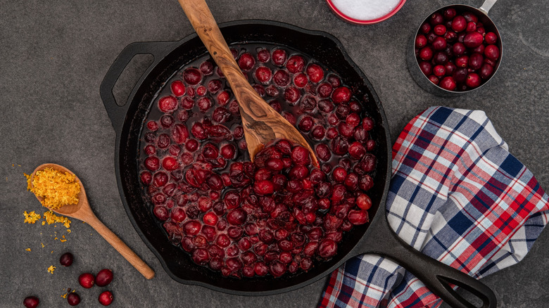 a cast iron skillet is filled with cranberry sauce and sits next to a spoonful of orange zest and fresh cranberries