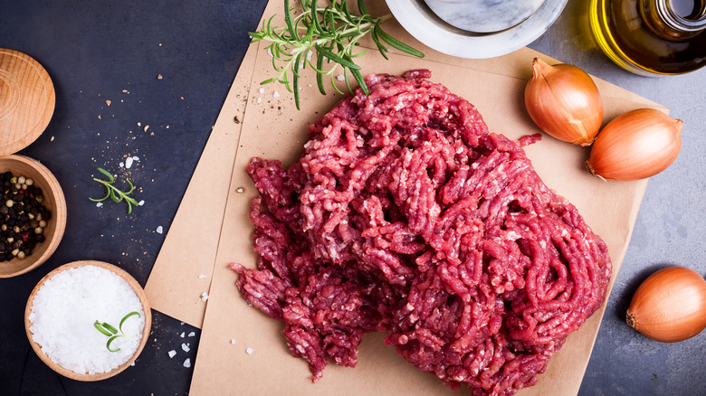 Ground beef sits in a mound on some parchment paper, surrounded by oil, salt, pepper, onions, and fresh rosemary