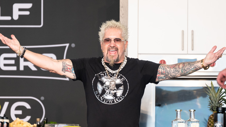 Smiling Guy Fieri in a black T-shirt with his arms spread wide.