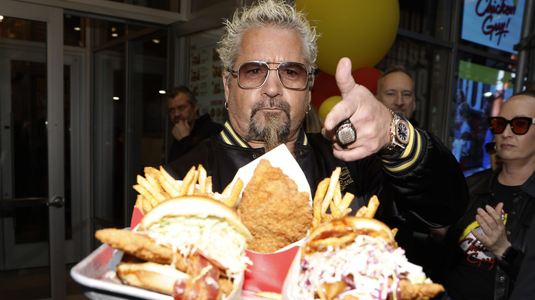 Guy Fieri holds a tray containing fried chicken, french fries, and chicken sandwiches at one of his Chicken Guy! restaurants