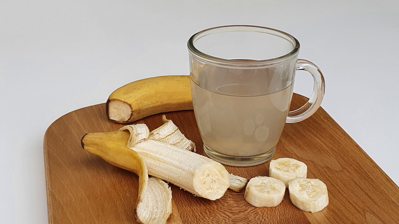Banana peel tea in a clear mug, with two bananas around. One banana is partially peeled and sliced. The other banana is still unpeeled, with top cut off. All on a wooden cutting board and a plain white background.
