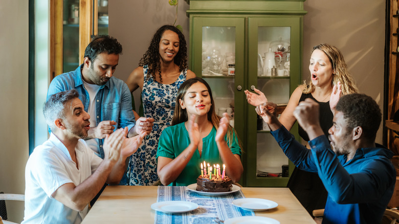 A group of friends gather around a birthday cake with candles, singing and clapping