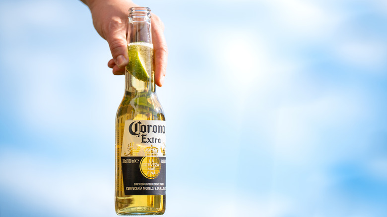 Bottle of Corona held in a hand against a blue-sky background.