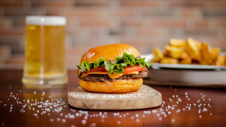 A smash burger on a wooden slab with a glass of beer and french fries in the background