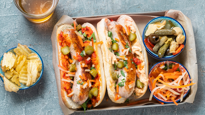 Two bratwurst sausages dressed as hot dogs, with lots of toppings and a side of chips and pickled vegetables